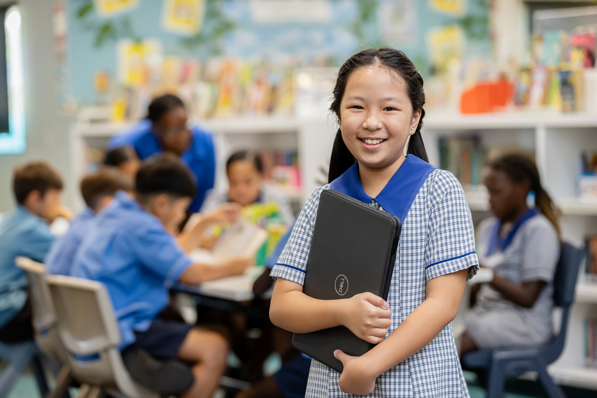 A smiling student standing in a classroom and holding a laptop. There are a group of students seated behind her.
