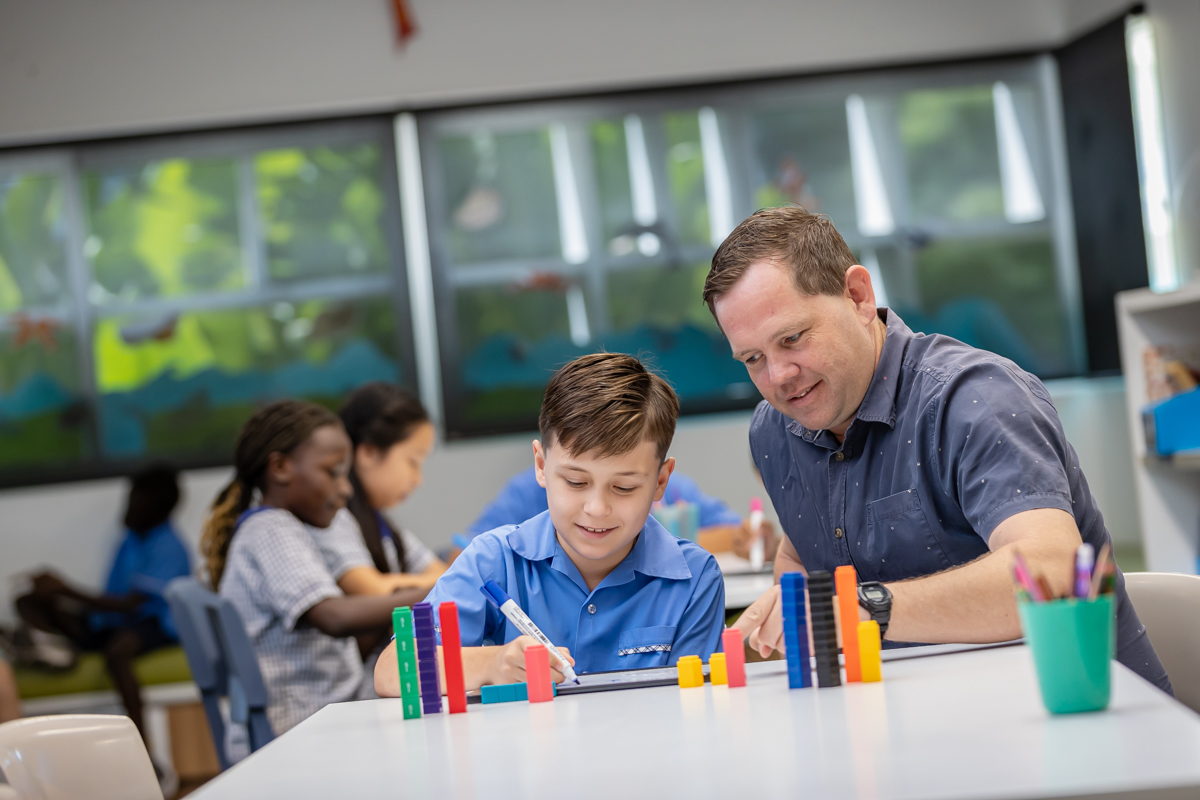 A teacher is sitting at a desk with a student. There is a group of students at a desk in the background.
